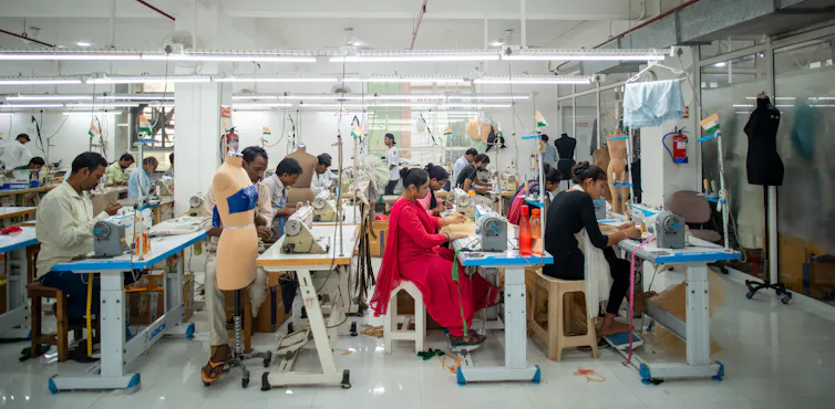 male and female indian garment producers sitting working at their sewing machines in a factory.