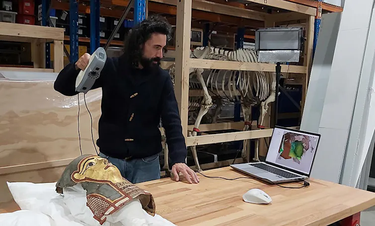 man, looking toward a laptop screen, holds small machine above an Egyptian mummy mask