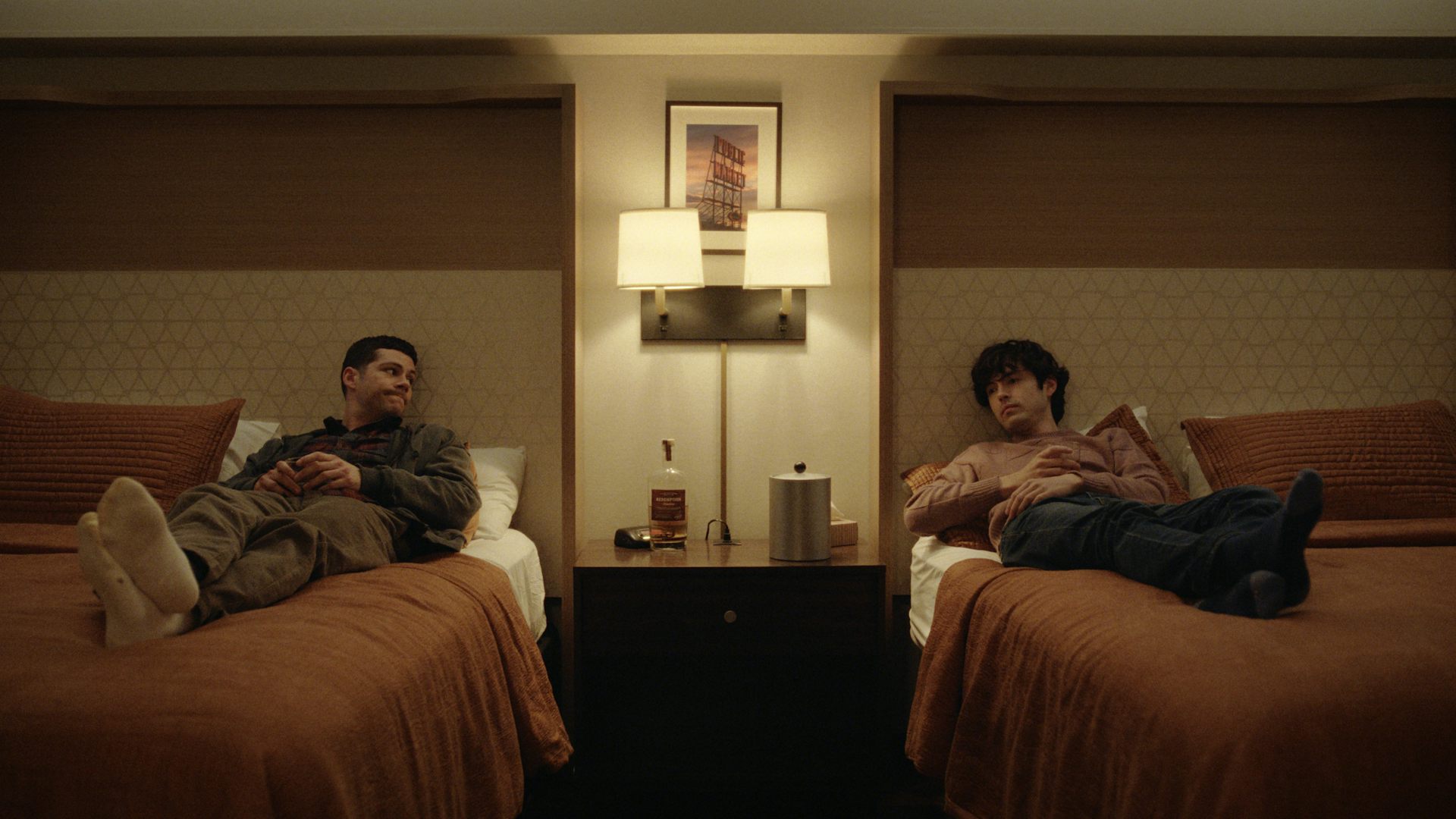 Two men in a twin hotel room each lying on a single bed looking at each other.