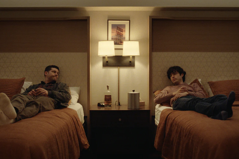 Two men in a twin hotel room each lying on a single bed looking at each other.