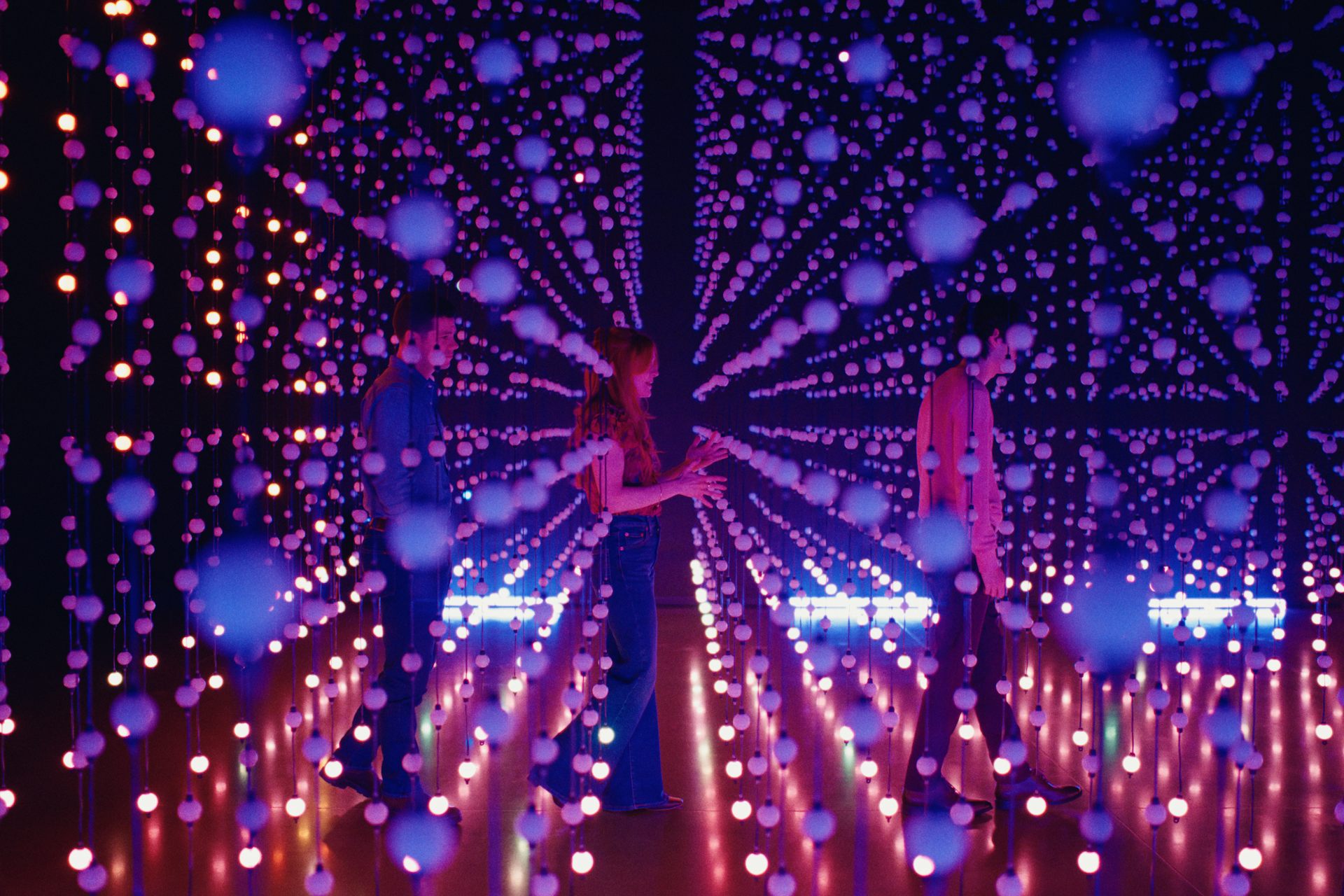 Two young men and a young woman on a mirrored dancefloor covered with lights.