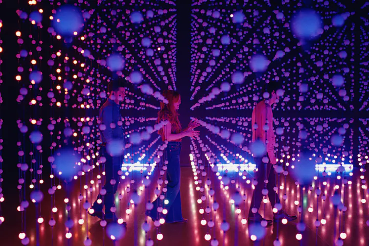 Two young men and a young woman on a mirrored dancefloor covered with lights.