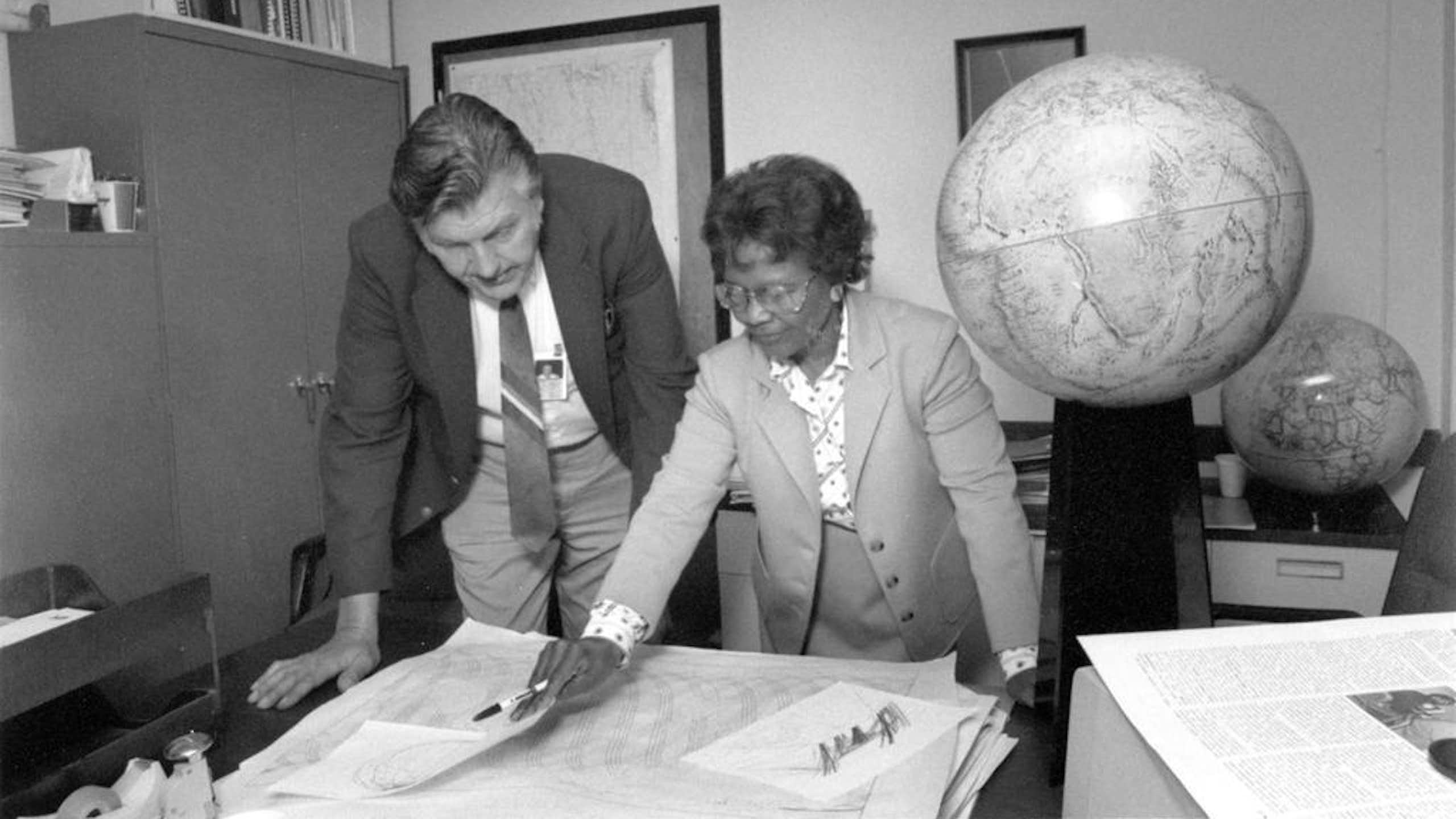Fotografia em preto e branco de uma mulher negra virando um documento sobre uma mesa ao lado de um homem branco, com um globo atrás deles.