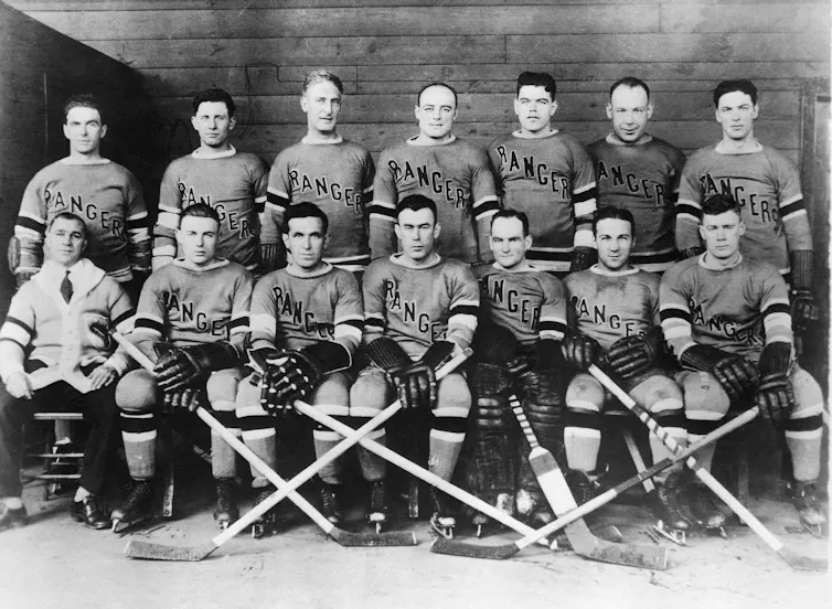 14 men in hockey uniforms, posed in two rows for a photo, some with their hockey sticks.