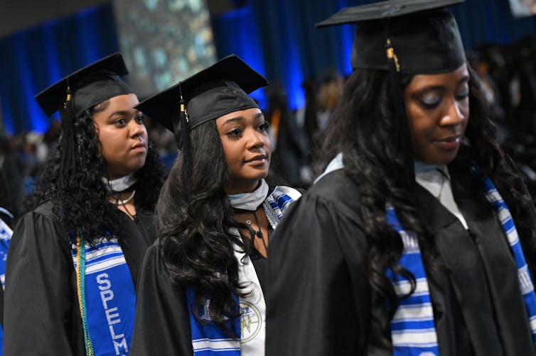 Three young women wear black graduation robes and black graduation hats and stand in a row.
