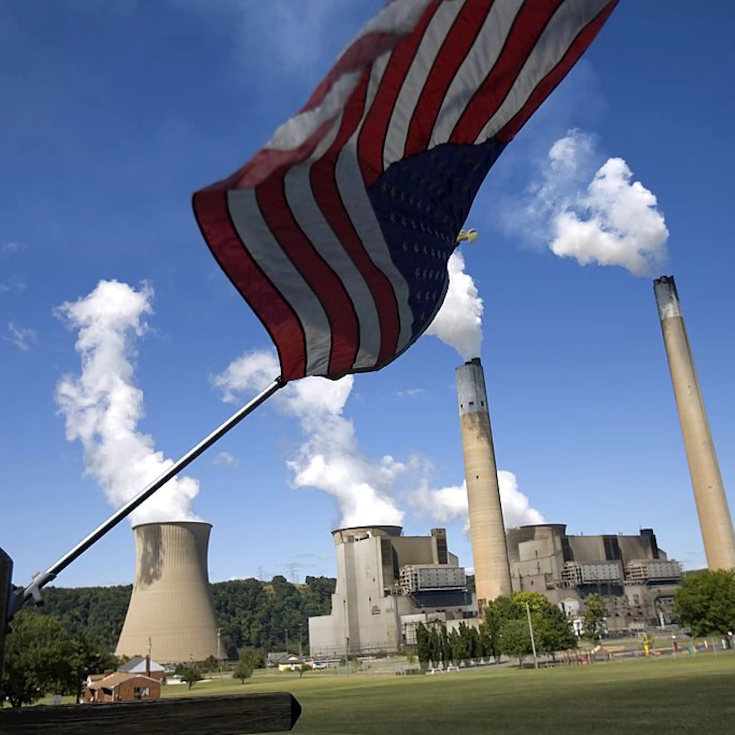 Smoke puffs out of a power plant with an American flag in the foreground of the photo.
