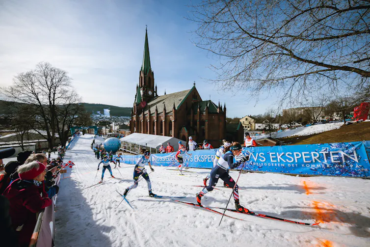 Female skiers race through a town with a church beside them, fans along the track and lots of snow-free ground outside the snowy race course.
