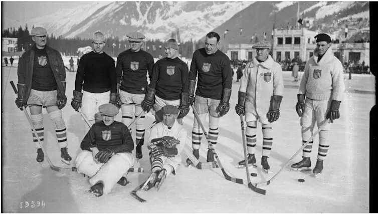 A group of men standing on snow in front of mountains, some holding hockey sticks.