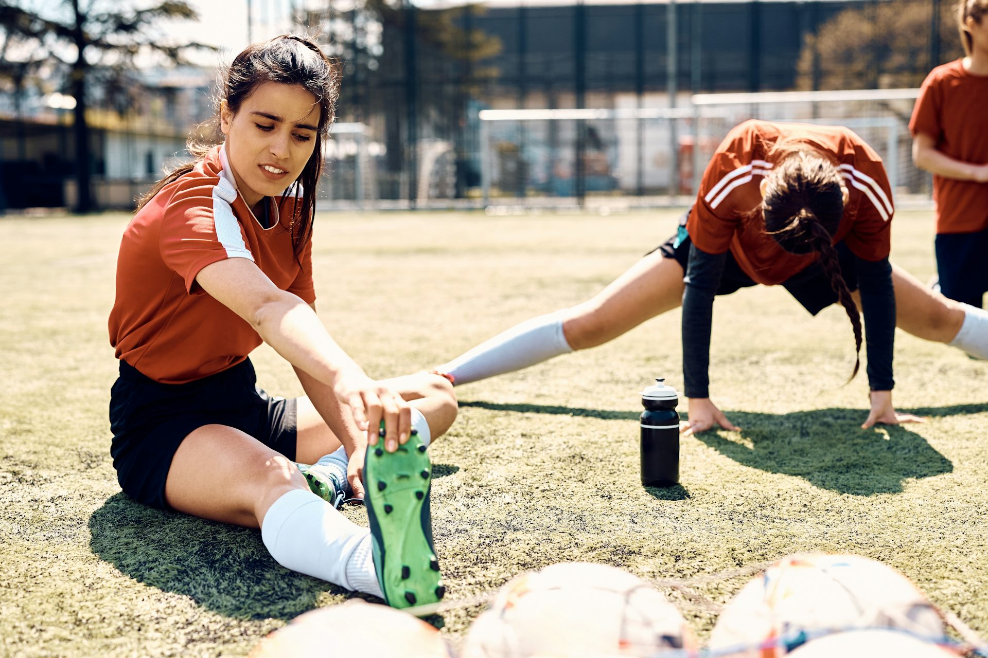 Dues joves amb equipació esportiva fan estiraments a terra en un camp de futbol.