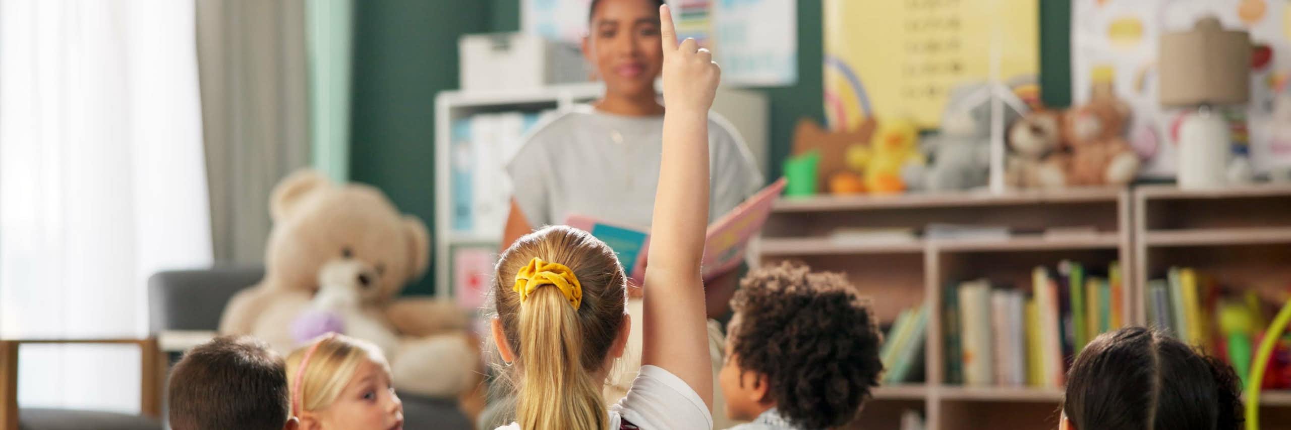 Una nena alça la mà en classe. Al seu voltant, més xiquets i una mestra mirant-la.