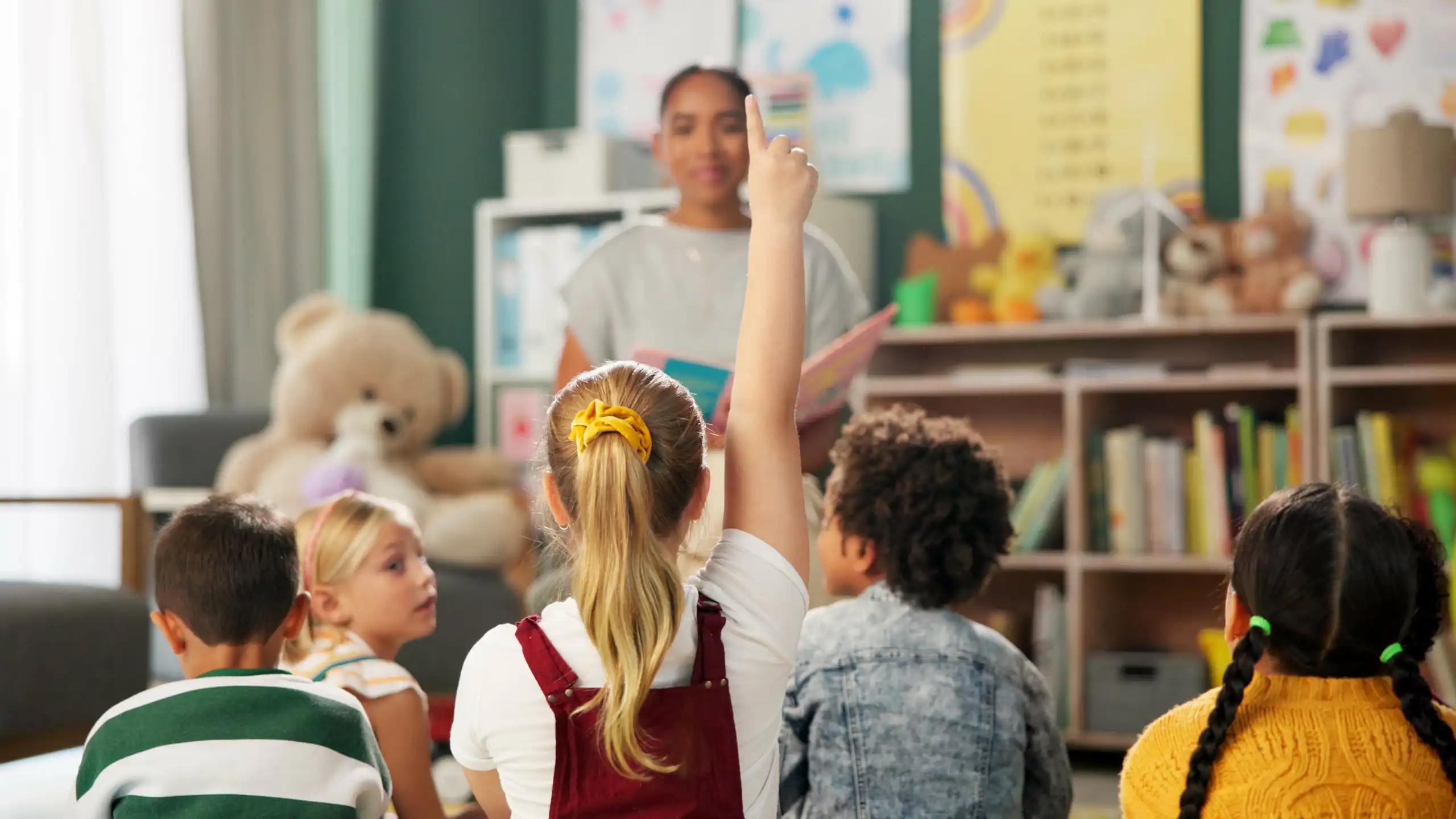 Una nena alça la mà en classe. Al seu voltant, més xiquets i una mestra mirant-la.