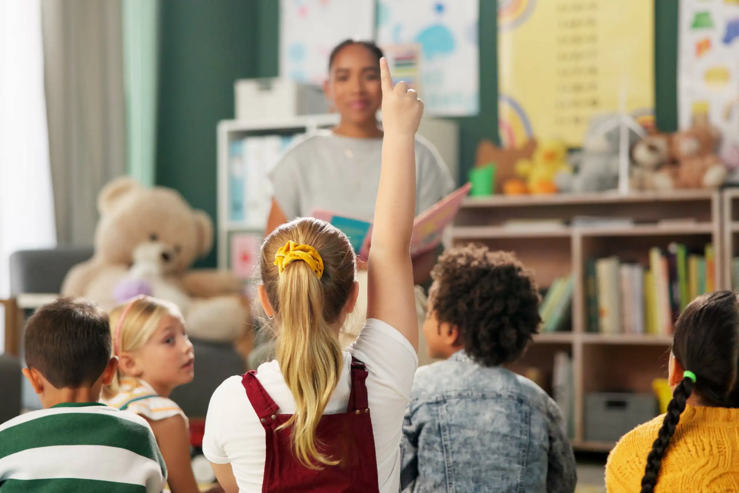 Una nena alça la mà en classe. Al seu voltant, més xiquets i una mestra mirant-la.