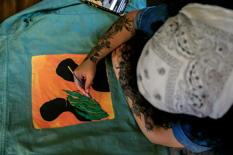 A Black painter's hand seen holding a brush painting two black silhouettes against an orange sky.