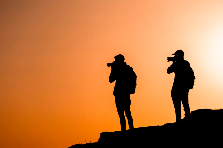 two people standing on hill holding cameras, silhouette black against orange sky