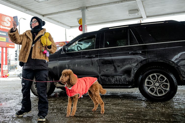 A woman wearing a yellow jacket holds her phone to record ICE agents in one hand and her dog's leash in the other.