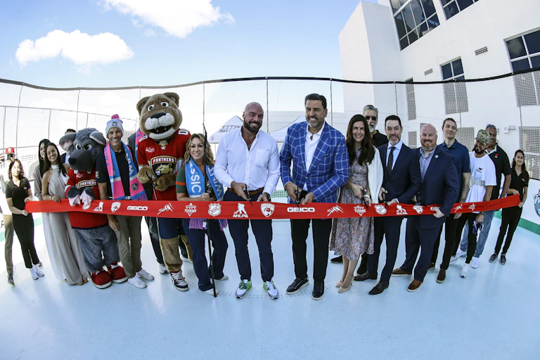 A group of people and costumed mascots prepare to cut a ribbon to open an ice skating rink.