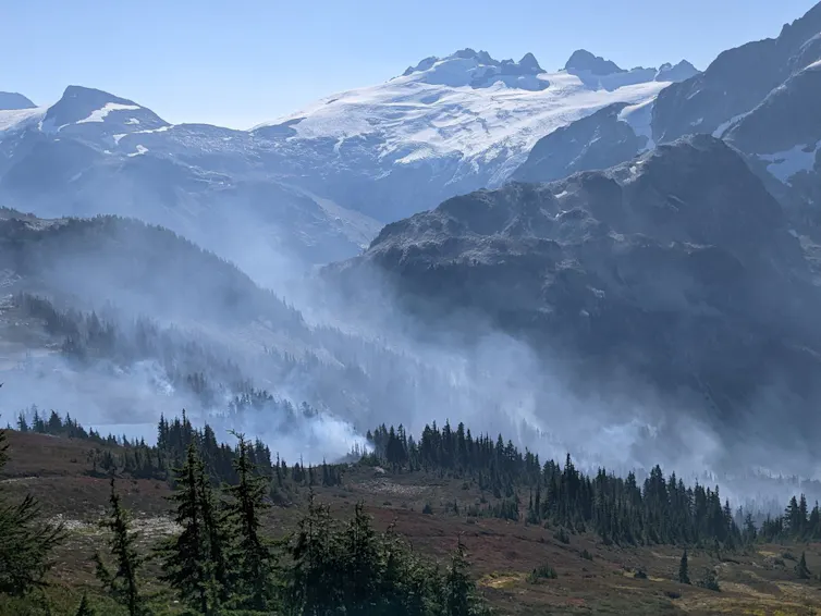 Smoke climbs above a wooded mountainside, with higher peaks in the background.