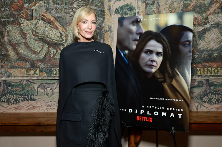 A sandy haired woman in a black coatdress, looking serious, next to a poster for a series called 'The Diplomat.'