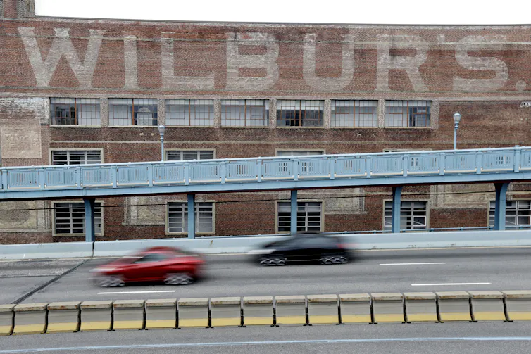 Cars on highway pass a large brick building with faded paint that reads 'Wilbur's'