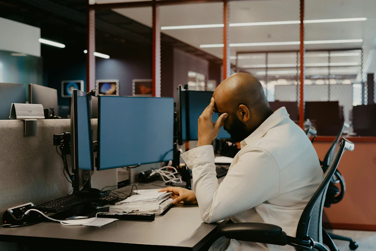 A man sitting at a desk rests his forehead against his hand