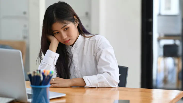 A young woman sits at a desk, resting her cheek against her hand