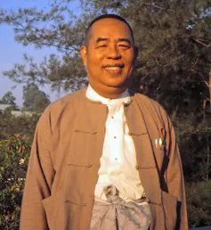 A man wearing a brown jacket smiles while standing outdoors, with trees in the background.