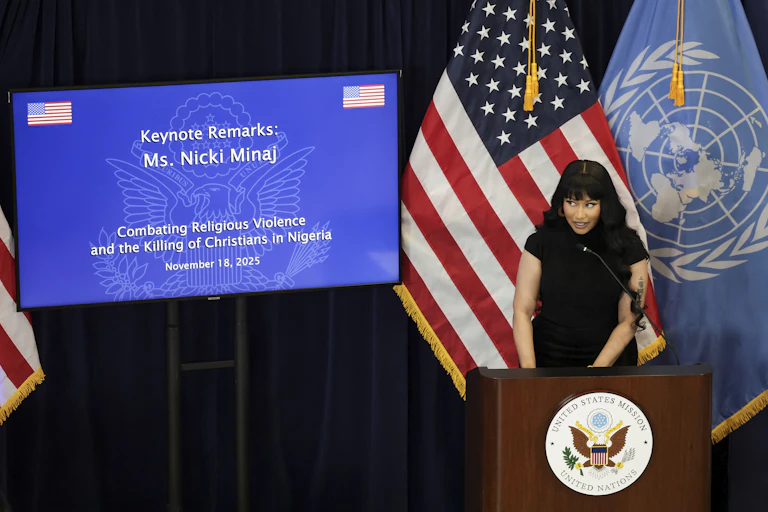 A woman in a black dress standing on a podium speaking into a micorphone with the US flag and a bright screen behind her