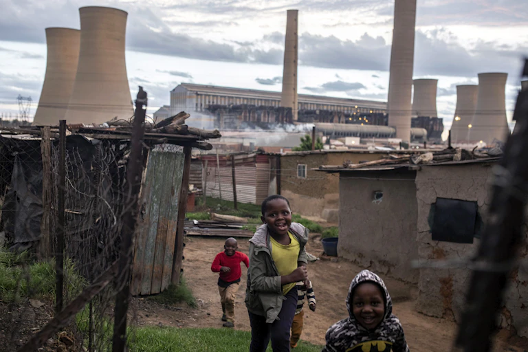 Children play among shacks, with power station cooling towers in the background
