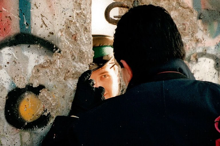 Two men speak through a crack in the wall