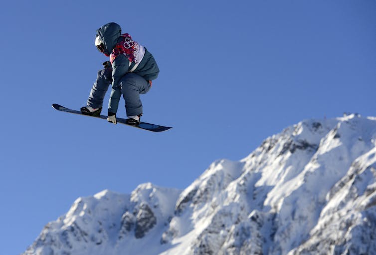 A snowboarder wearing a puffy jacket, snow pants and an Olympic rings bib, grips his board as he flies through the sky.