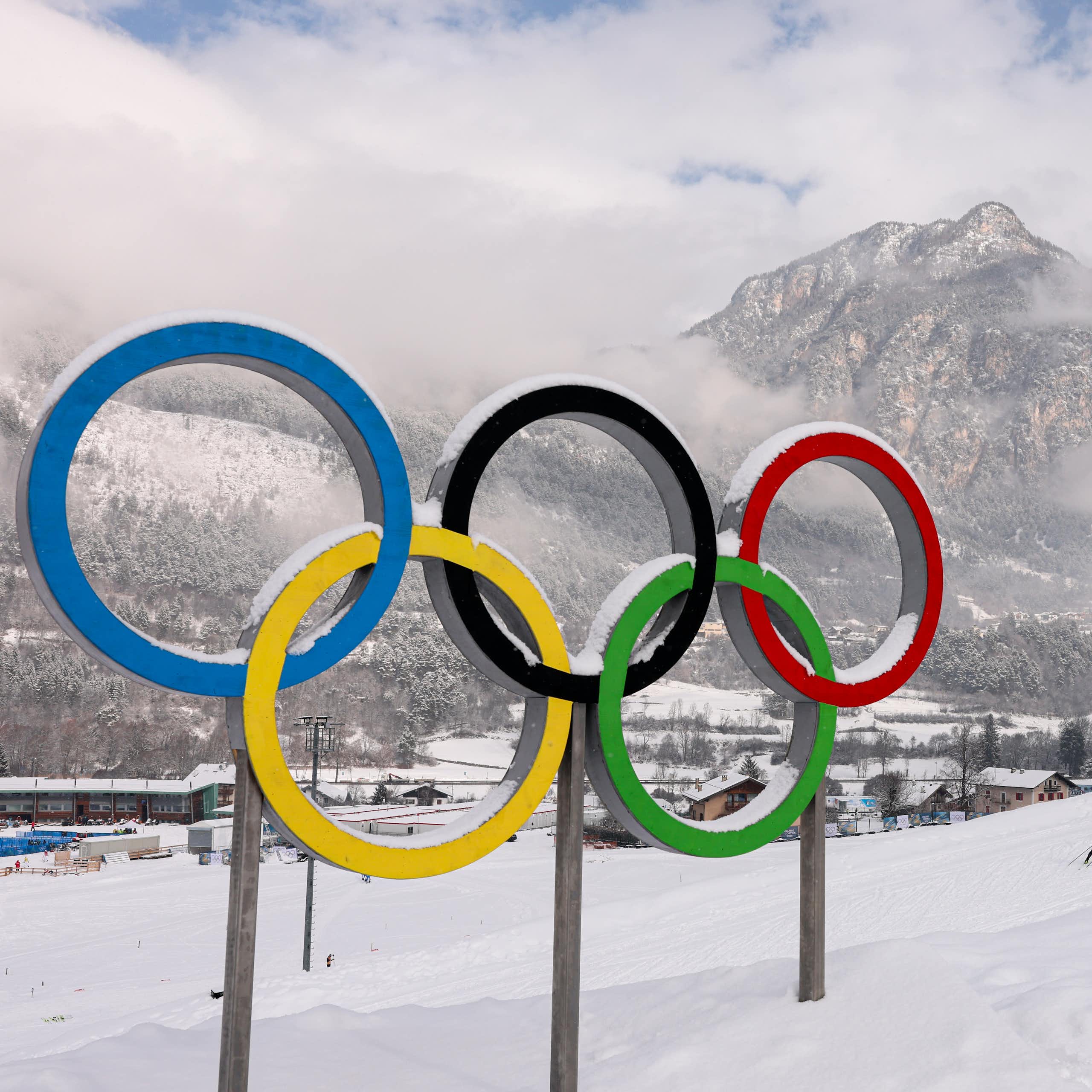 A statue of the Olympic Rings covered in a dusting of snow, with a mountain and a cross-country ski track in the background.