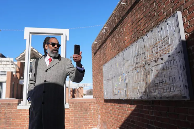 Man in overcoat and sunglasses holds up phone, with brick walls around him