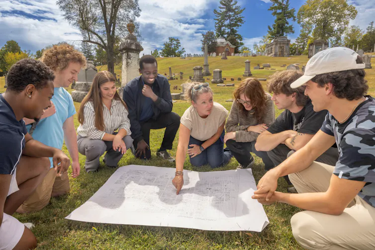 Group of people surrounding a map laid on a grassy field, headstones and trees in the background