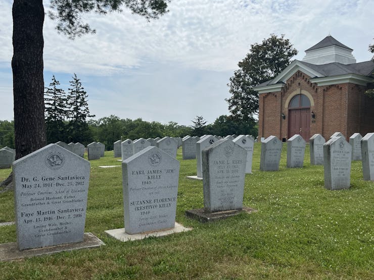 Rows of headstones in a cemetery, trees and a brick building in the backdrop