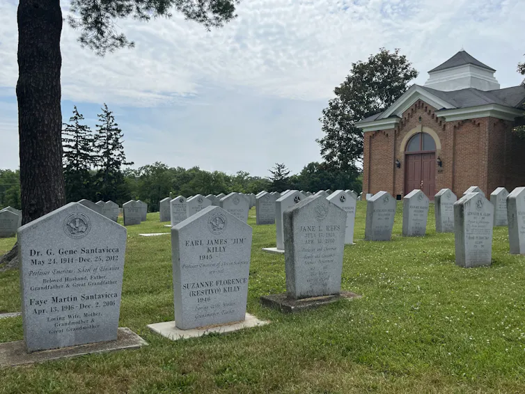 Rows of headstones in a cemetery, trees and a brick building in the backdrop