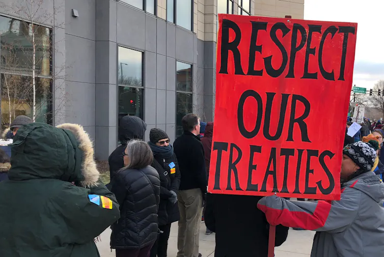 People gather outside a building; one is holding a sign saying 'Respect our treaties.'