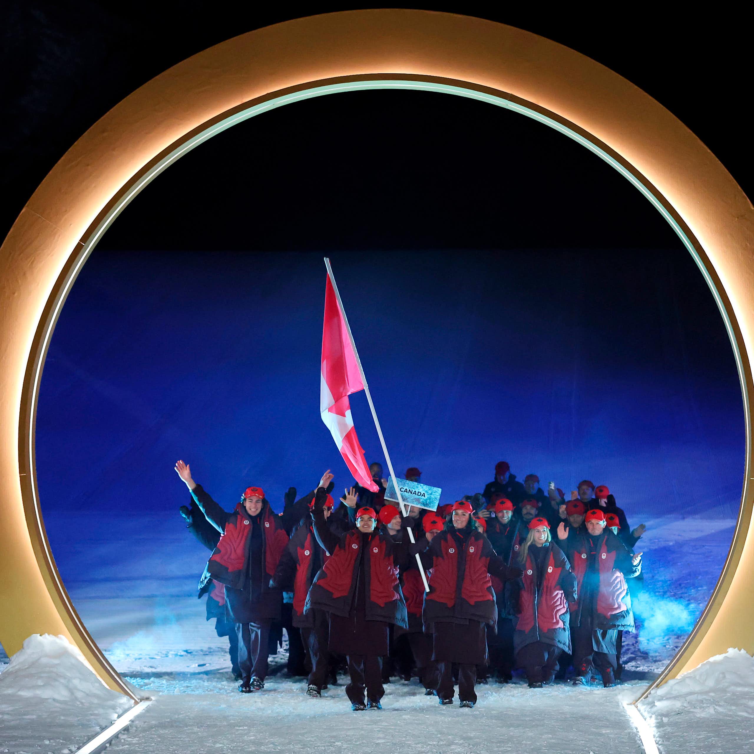A group of people in maroon jackets march through a giant gold arch behind someone waving a big Canadian flag