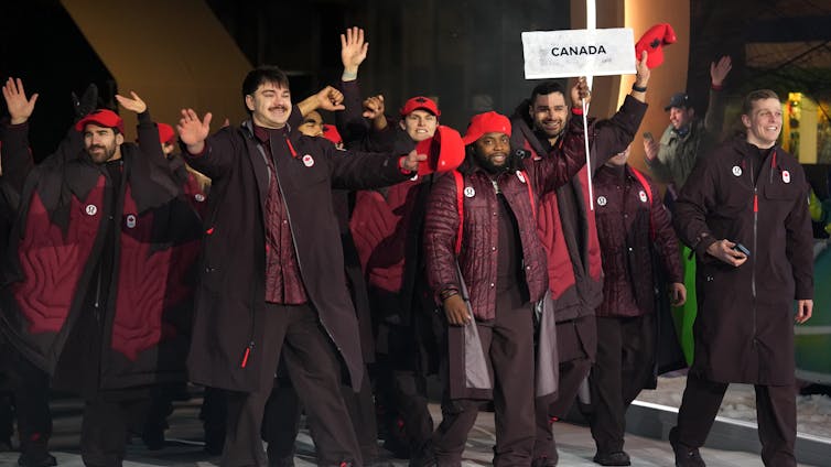 Cómo los atletas indígenas están desafiando ideas simples de unidad nacional en los Juegos Olímpicos 1 Un grupo de personas con chaquetas granates caminan detrás de alguien con el cartel Canadá.