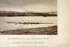 Black and white photo of four men rowing in a row boat across a body of water