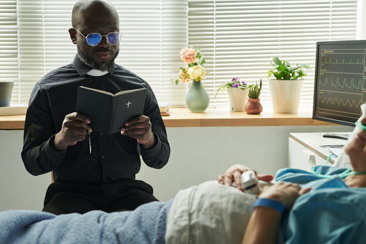 A bespectacled Black priest reads from the Bible at a patient's bedside in a hospital.