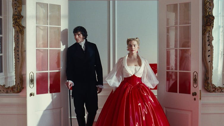 A tall, handsome, brooding man and a beautiful blonde woman in a red leatherette and blousy white gown