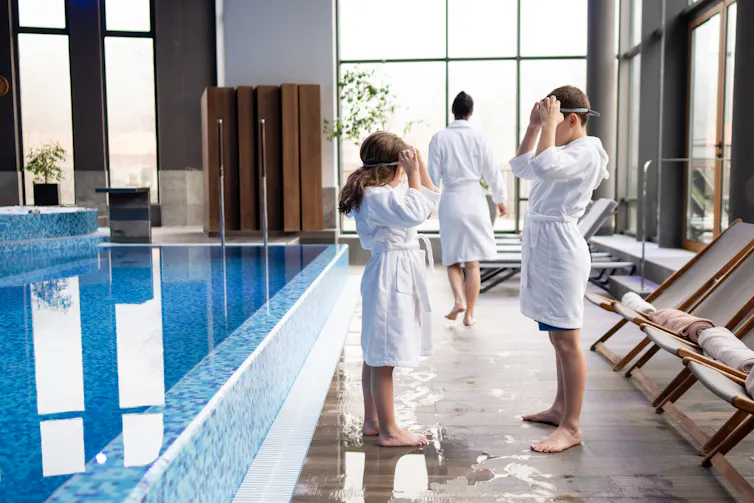 A young boy and girl in bath robes put on their goggles next to an indoor pool.