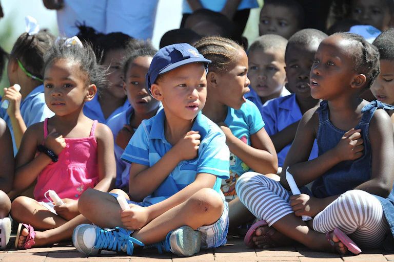 A group of children sitting cross-legged, with one fist on their chest