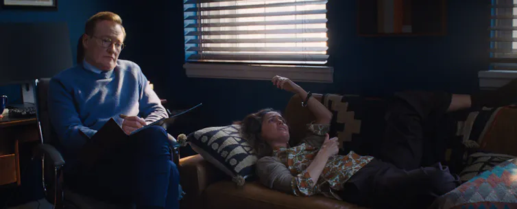 A man with red hair and glasses looks pained as a woman lies on a couch speaking.