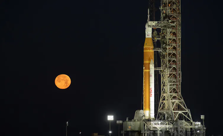 Una nave espacial y un cohete en una plataforma de lanzamiento por la noche con la Luna a la izquierda de la imagen