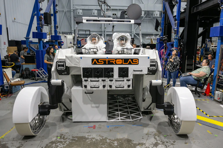 Dos personas con trajes de astronautas sentados en un vehículo lunar en el que pone "Astrolab" rodeados de operarios