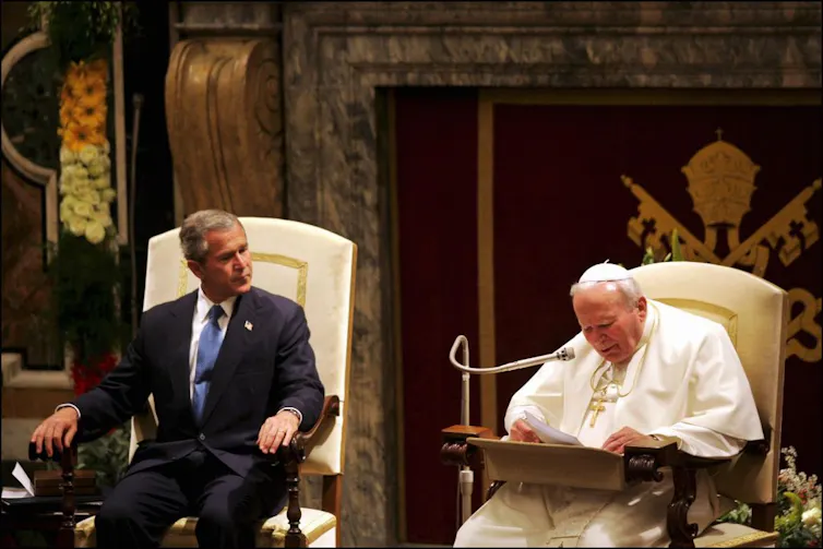 A man in a suit and blue tie sits in an ornate white chair, next to another seated man in white robes reading into a microphone.