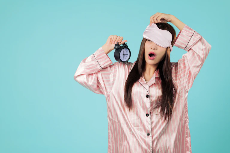 Woman in pink pyjamas and eye mask looks at alarm clock with surprised expression.