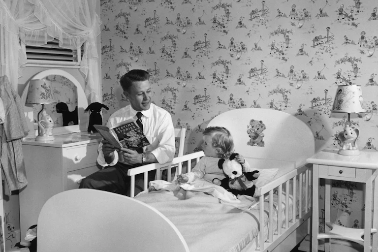 A man holds a book that says 'Bedtime stories' as he sits next to a child sitting up in bed in a black and white photo.