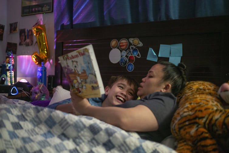 An East Asian woman lies on the bed next to a smiling boy and holds a book close to their faces.